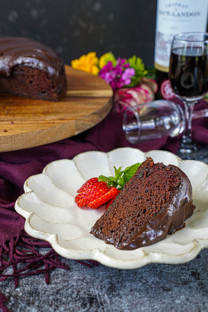 A slice of Red Wine Chocolate Cake on a plate with a glass and bottle of red wine in the background.