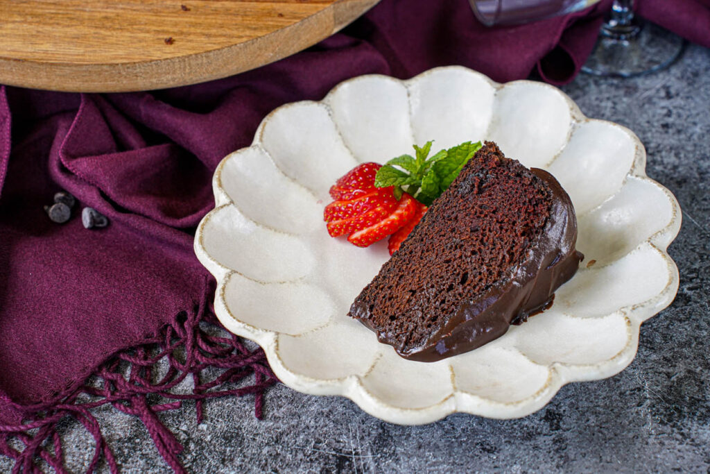 A slice of Red Wine Chocolate Cake on a plate with sliced strawberry and a mint sprig.