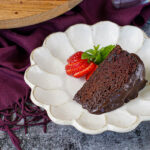 A slice of Red Wine Chocolate Cake on a plate with a sliced strawberry and mint sprig.