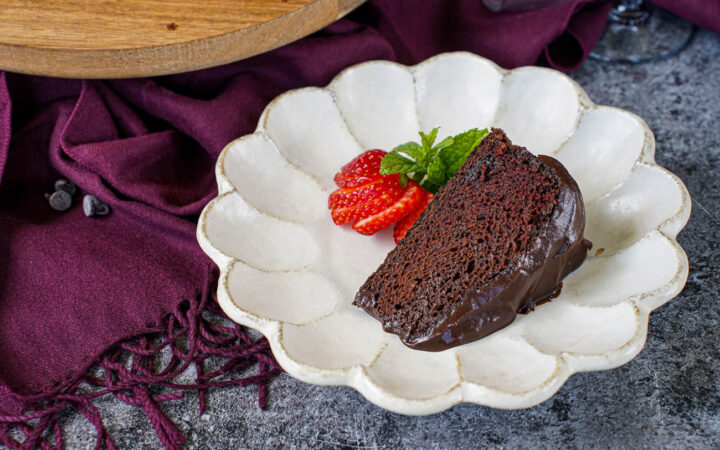 A slice of Red Wine Chocolate Cake on a plate with a sliced strawberry and mint sprig.