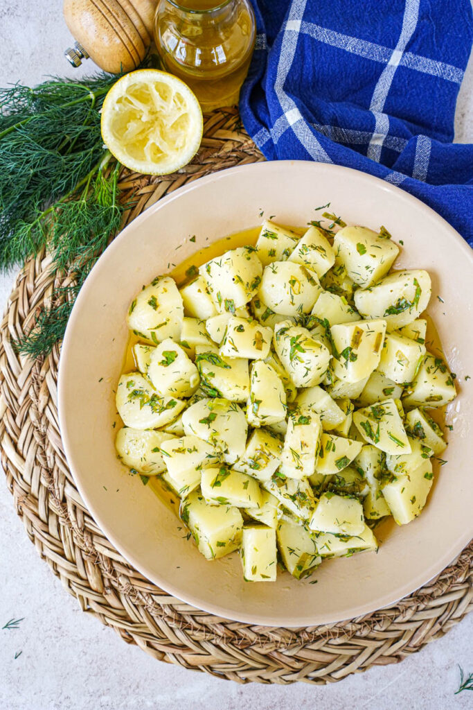 Aerial view of Patatosalata (Herby Potato Salad) in a bowl next to a lemon half, dill, and olive oil.