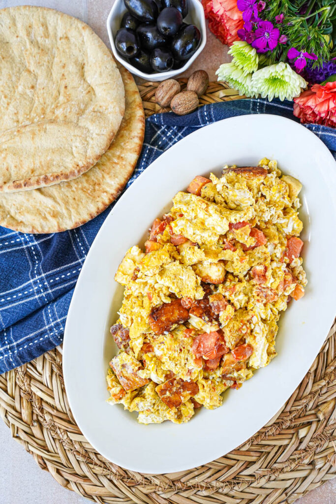 Aerial view of Scrambled Eggs with Halloumi on an oval white platter next to bread, olives, nutmeg, and flowers.
