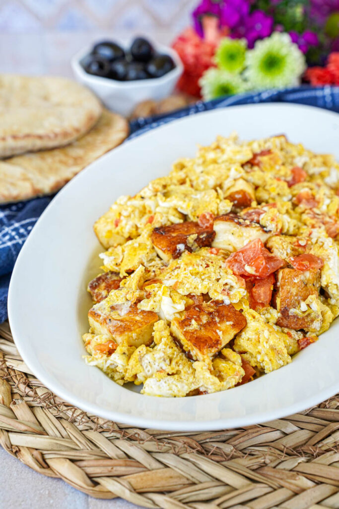 Side view of Scrambled Eggs with Halloumi on an oval white platter with bread, olives, and flowers in the background.