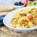 Scrambled Eggs with Halloumi on a white oval platter with bread and black olives in the background.