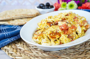 Scrambled Eggs with Halloumi on a white oval platter with bread and black olives in the background.