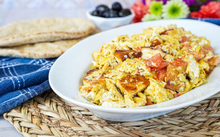 Scrambled Eggs with Halloumi on a white oval platter with bread and black olives in the background.