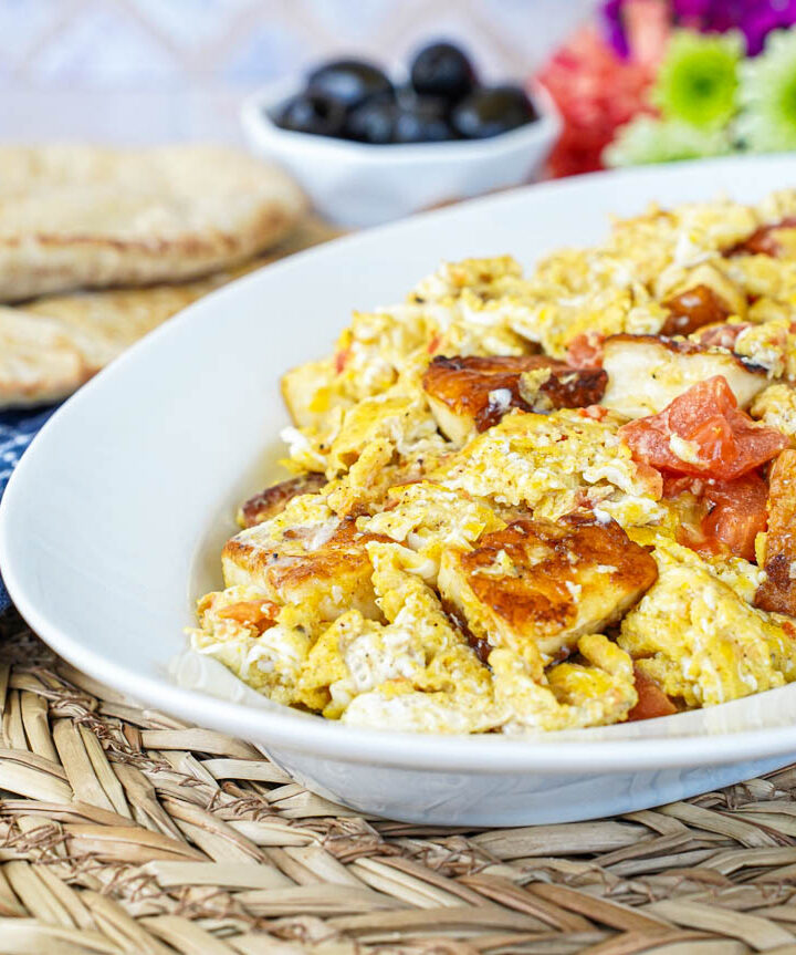 Scrambled Eggs with Halloumi on a white oval platter with bread and black olives in the background.