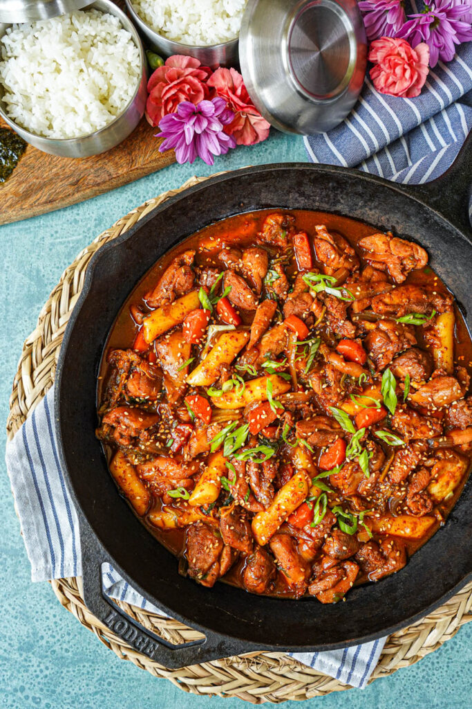 Aerial view of Dakgalbi (Spicy Stir-Fried Chicken) in a cast iron skillet next to bowls of rice and flowers.