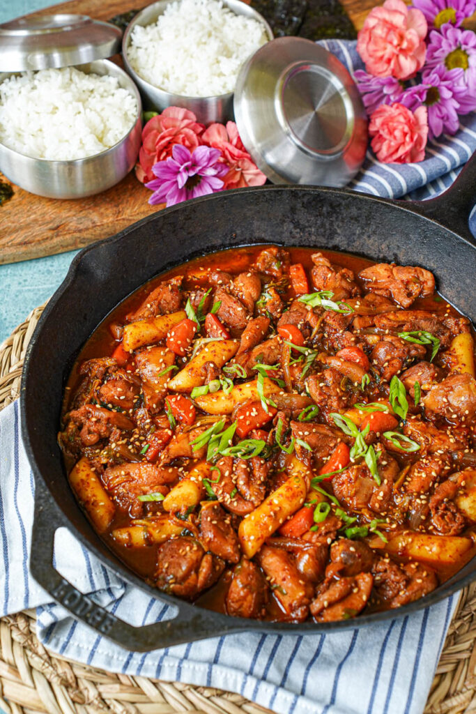 Close up of Dakgalbi (Spicy Stir-Fried Chicken) in a cast iron skillet with rice and flowers in the background.