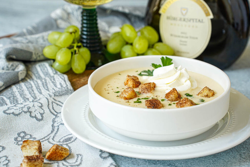 Fränkische Weinsuppe (Franconian Wine Soup) in a white bowl with a bottle of wine and green grapes in the background.