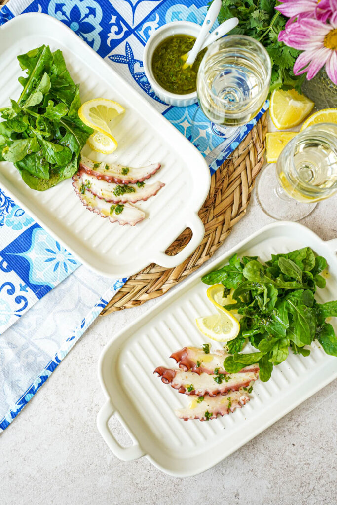 Aerial view of Grilled Octopus Carpaccio on two plate plates next to a bowl of cilantro lemon dressing and two glasses of wine.