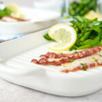 Three slices of Grilled Octopus Carpaccio on a white plate with arugula and a lemon slice.