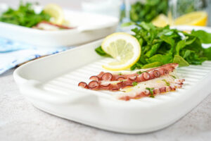 Three slices of Grilled Octopus Carpaccio on a white plate with arugula and a lemon slice.