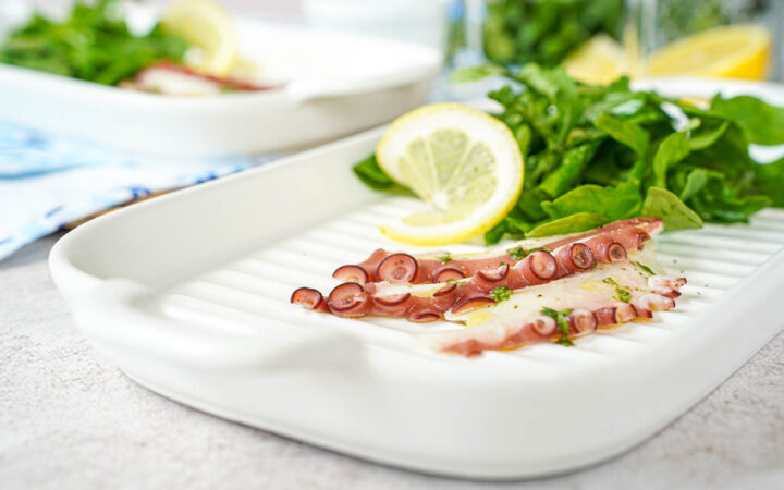 Three slices of Grilled Octopus Carpaccio on a white plate with arugula and a lemon slice.