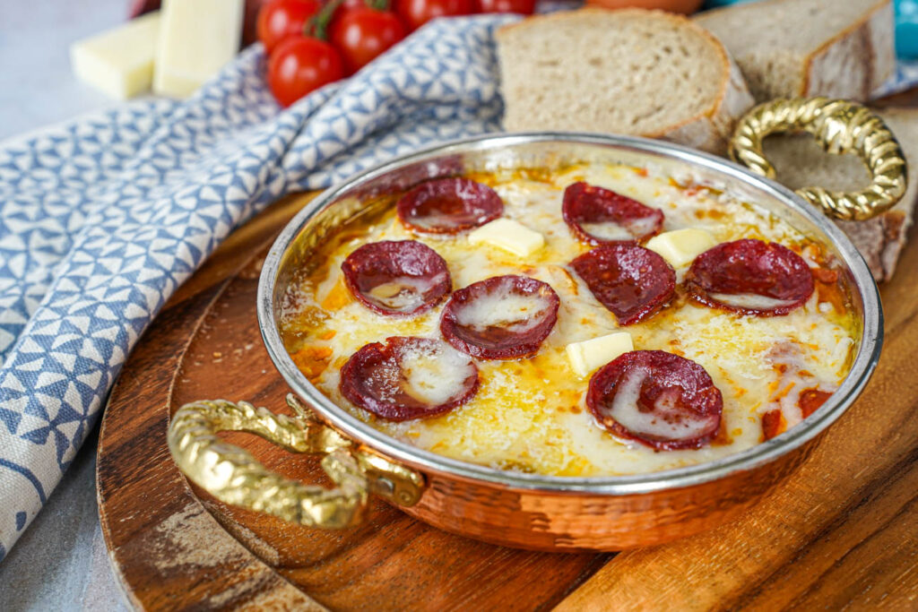Menemen (Tomato and Pepper Scrambled Eggs) in a copper bowl with melted cheese and sucuk slices.