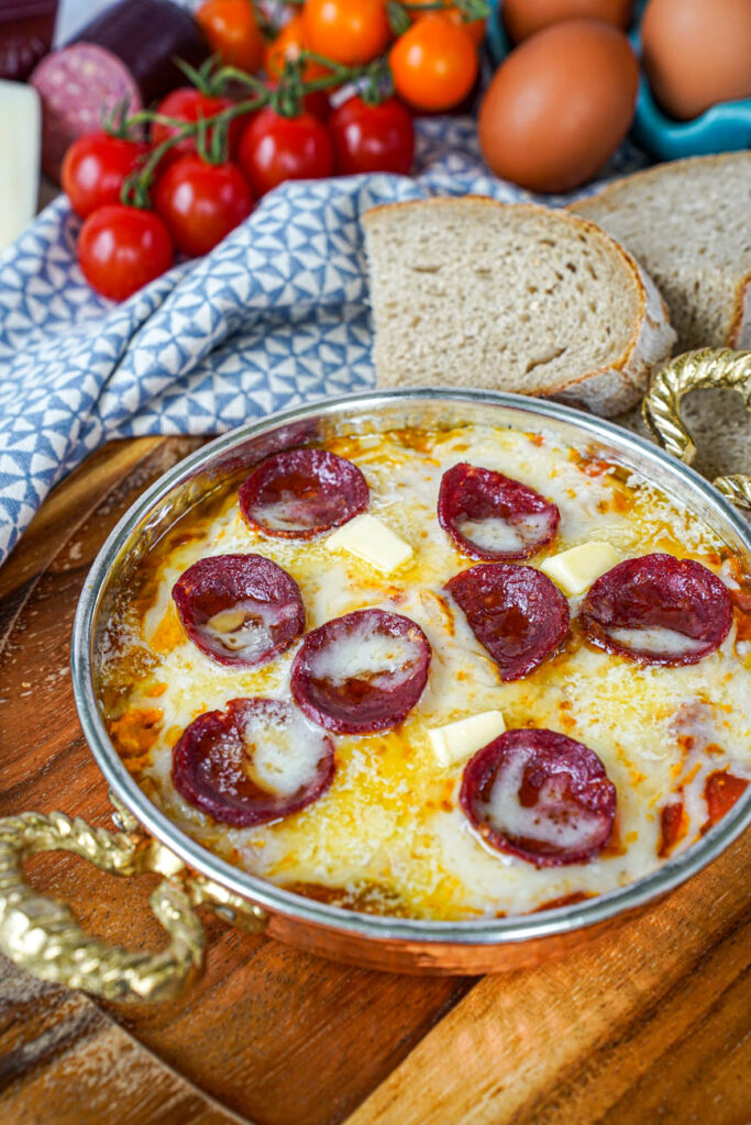 Close up of Menemen (Tomato and Pepper Scrambled Eggs) in a copper bowl with bread, eggs, and cherry tomatoes in the background.