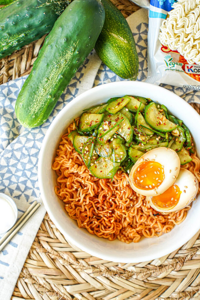 Aerial view of Korean Cucumber Salad Ramyeon in a bowl next to cucumbers and instant noodle packets.