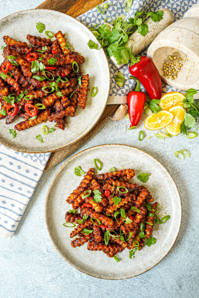 Aerial view of Masala Fries on two plates next to red chilies, lemon wedges, coriander, garlic cloves, and cilantro.