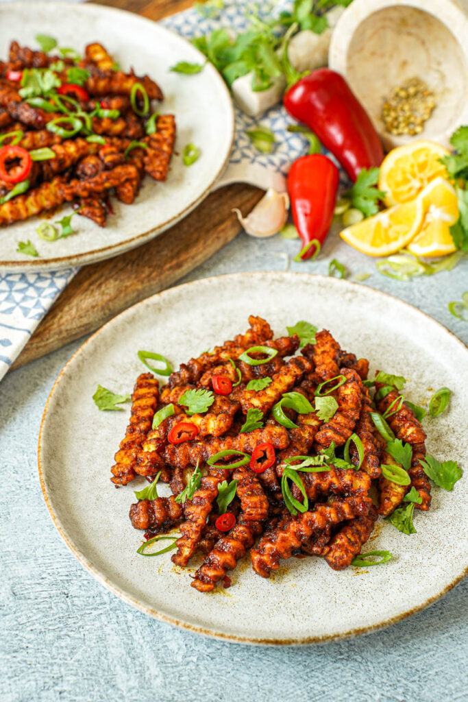 Close up of Masala Fries with red chilies, garlic cloves, lemons, and cilantro in the background next to another plate.