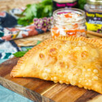 Two Pate Kòde (Haitian Fried Patties) on a wooden board with a jar of pickled cabbage.