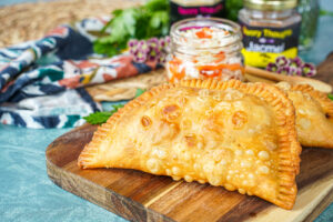 Two Pate Kòde (Haitian Fried Patties) on a wooden board with a jar of pickled cabbage.