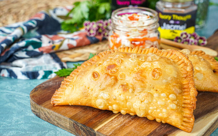 Two Pate Kòde (Haitian Fried Patties) on a wooden board with a jar of pickled cabbage.