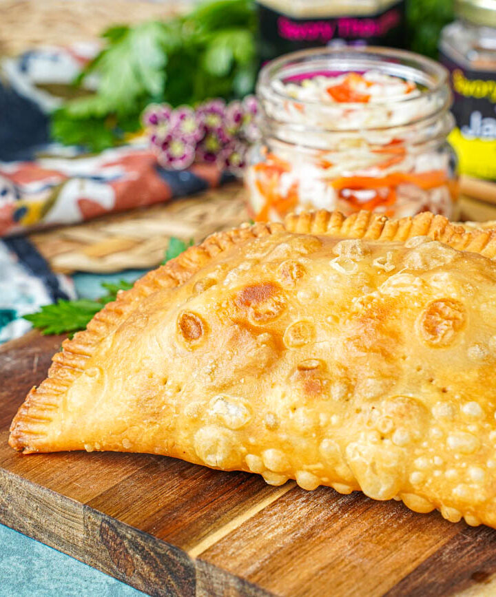 Two Pate Kòde (Haitian Fried Patties) on a wooden board with a jar of pickled cabbage.