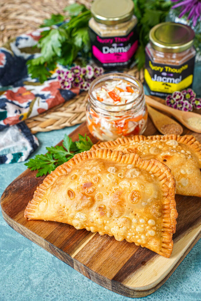 Pate Kòde (Haitian Fried Patties) on a wooden board next to a jar of pickled cabbage and two jars of spice blends.