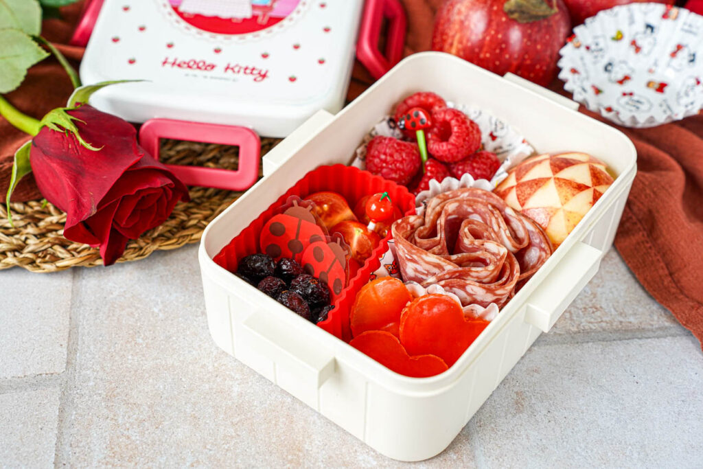 Red Bento Box theme with dried cranberries, halved cherry tomatoes, raspberries, heart-shaped bell peppers, salami, and an apple with a triangle design.