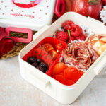 Red Bento Box lunch of heart-shaped bell peppers, salami, triangle design on an apple half, dried cranberries, cherry tomatoes, and raspberries.