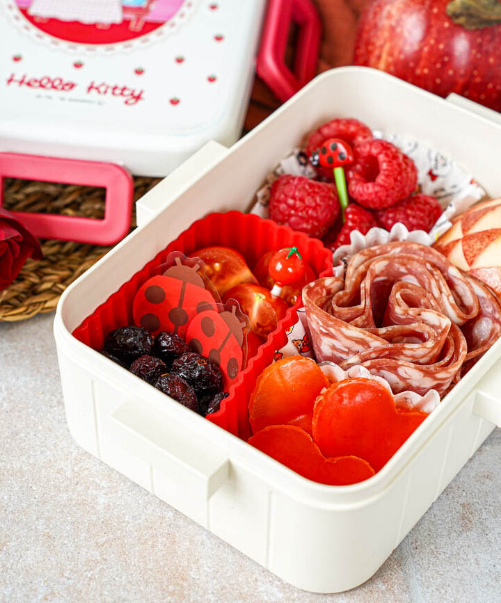 Red Bento Box lunch of heart-shaped bell peppers, salami, triangle design on an apple half, dried cranberries, cherry tomatoes, and raspberries.