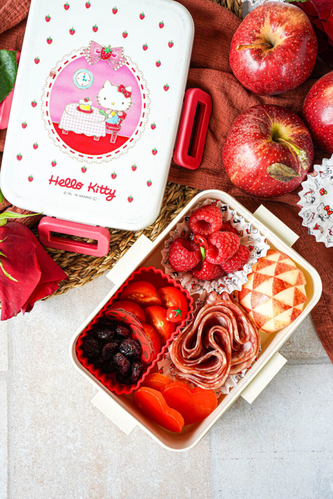 Aerial view of Red Bento Box next to apples, a red rose, and a Hello Kitty bento cover.