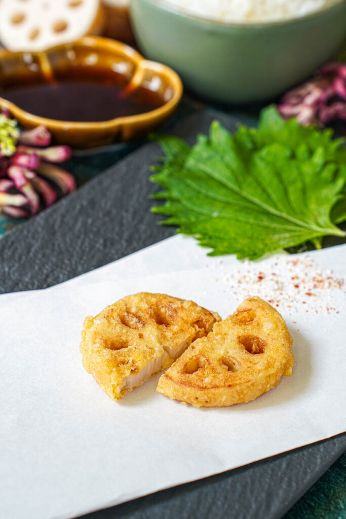 Two slices of Lotus Root Tempura on a folded white sheet of paper with shiso leaves, purple flowers, and dipping sauce in a brown bowl in the background.