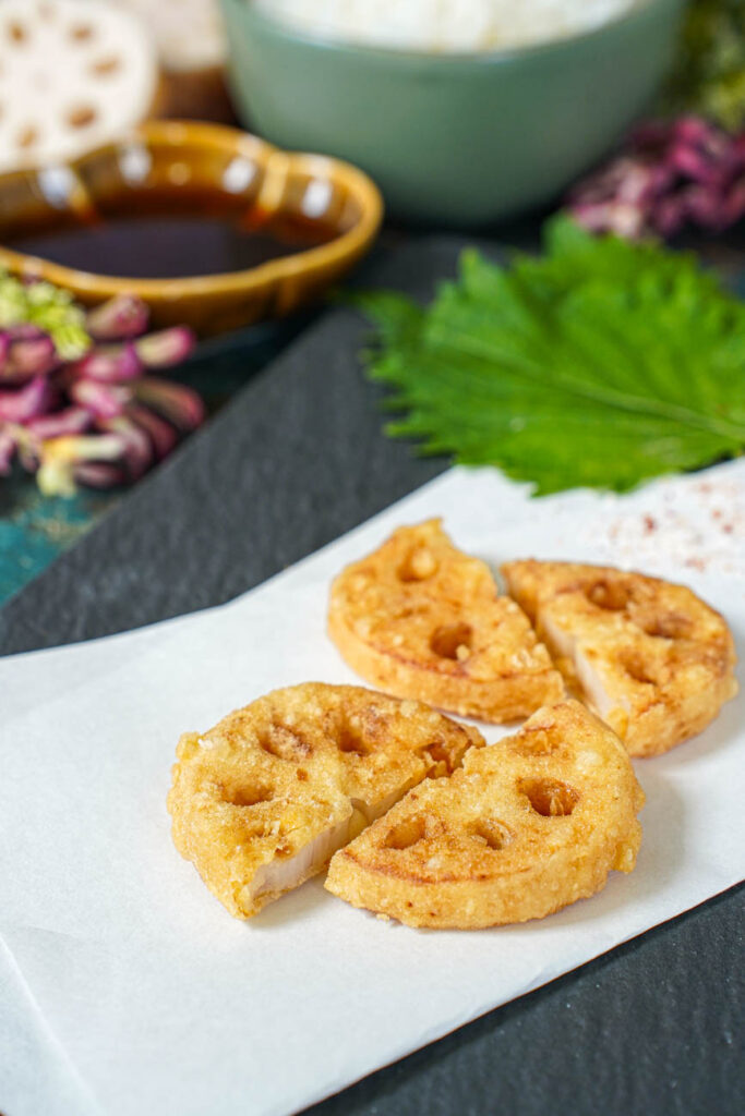 Four Lotus Root Tempura slices on a white folded sheet of paper with shiso leaves and purple flowers in the background.