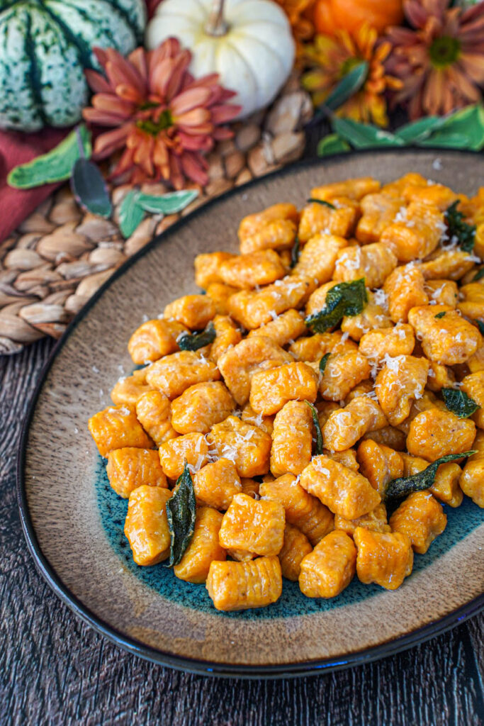 Close up of Pumpkin Ricotta Gnocchi on an oval plate with sage leaves, flowers, and pumpkins in the background.