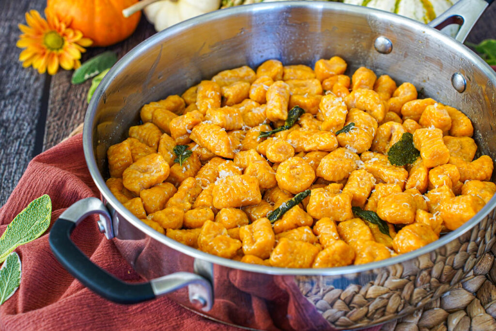 Pumpkin Ricotta Gnocchi in a steel saucepan next to sage leaves, yellow flowers, and pumpkins.