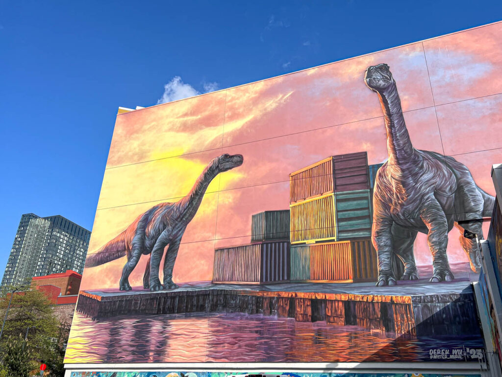 Mural on a wall of two long necked dinosaurs next to shipping containers.