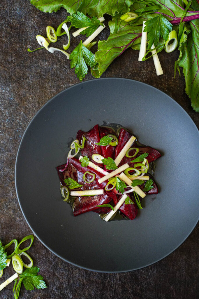 Aerial view of Tangy Beetroot Salad in a black bowl next to beet leaves and scallions.