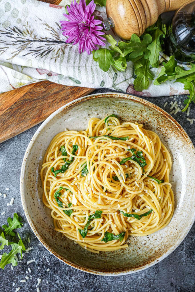 Aerial view of Spaghetti with Balsamic Butter in a tan bowl next to arugula leaves, a jar of balsamic vinegar, and purple flowers.