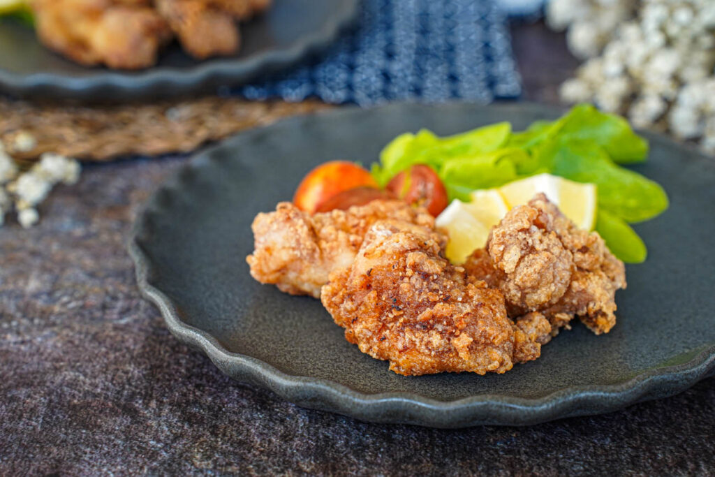 Three Tatsutaage (Marinated Fried Chicken) pieces on a grey plate with lettuce, cherry tomatoes, and lemon wedges.